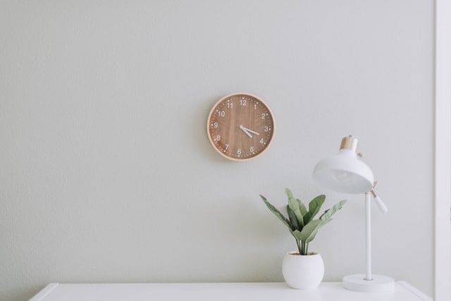 Picture of clock on a wall, and a desk with a plant and a lamp on it.