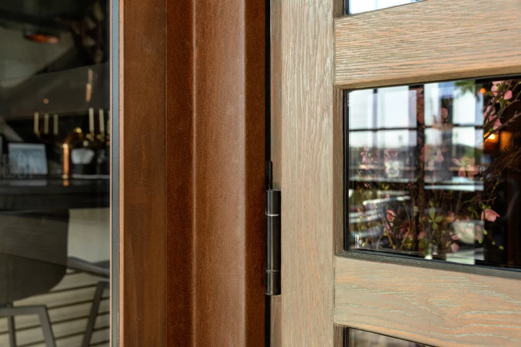 Detailed view of a door, featuring a warm reddish-brown corten steel frame with a smooth, polished finish, contrasting against a lighter-toned wood section with visible grain, accompanied by a sleek, black hinge and reflecting an inviting interior space with floral accents.