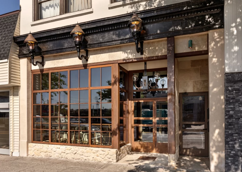 Exterior perspective of a modernized building with large corten steel windows divided into a grid pattern, a matching wooden door with glass panels to the right, a stone base along the facade, and decorative lantern-style lighting fixtures mounted above, casting shadows under a black cornice and surrounded by a neutral-toned exterior.